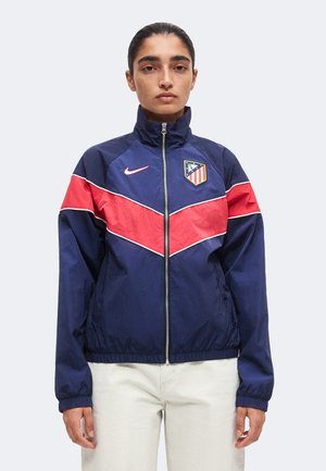 Personne aux cheveux foncés portant une veste de sport bleu marine et rouge zippée avec un pantalon blanc, debout devant un fond clair uni.