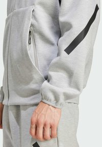 Šedý športový sako so zipsovou kapsou, s čiernym pruhom na rukáve a rebrovanými manžetami, vyrobené z mäkkej, textúrovanej látky.