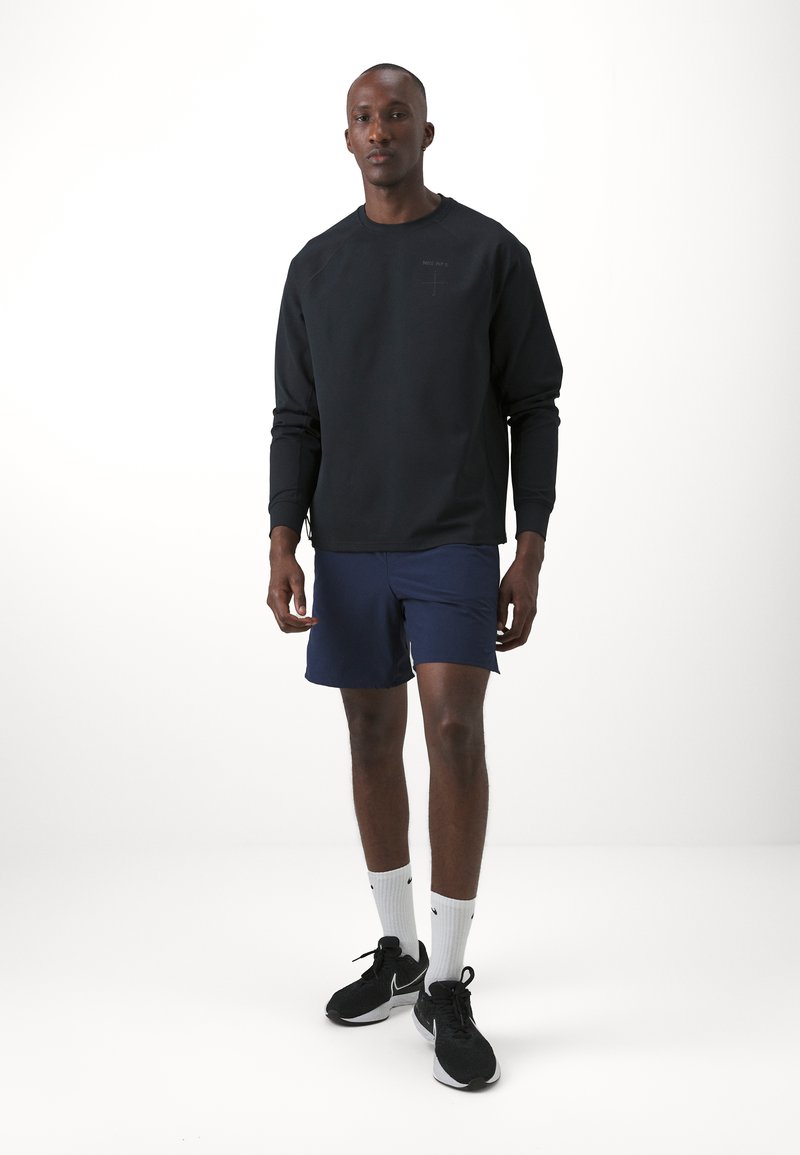 Sudadera negra con mangas largas y cuello redondo, combinada con pantalones cortos azul marino. El modelo lleva zapatillas negras y calcetines blancos. Diseño minimalista.