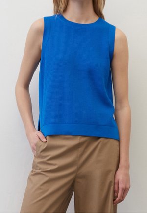 Haut en maille bleu texturé sans manches porté avec un pantalon marron clair, main gauche du modèle dans la poche, sur un fond clair uni.