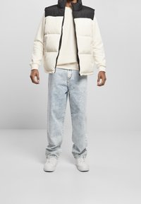Beige und schwarze wattierte Weste, getragen über einem cremefarbenen Sweatshirt. Hellblaue, ausgebleichte Jeans mit lockerer Passform und weißen Sneakers.