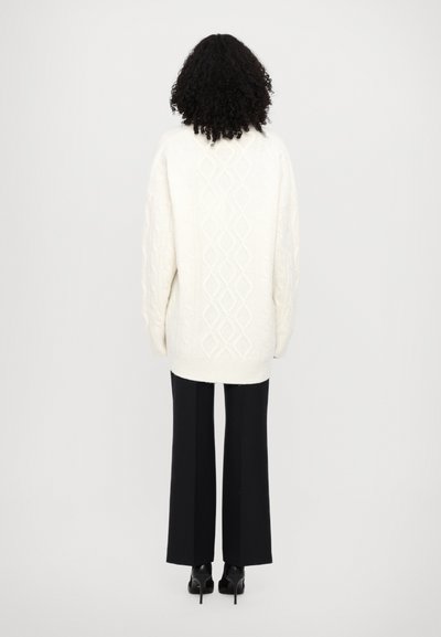 Pull en tricot blanc à mailles torsadées, avec une coupe ample, présentant un motif en losange. Associé à un pantalon large noir et des chaussures à talons noires.