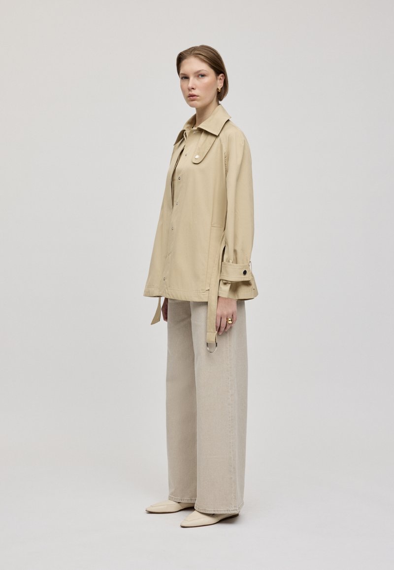 Beige Knopfmantel mit Kragen und seitlichen Taschen, kombiniert mit hellbeigen weit geschnittenen Jeans und spitzen Ballerinas, minimalistisches Design.