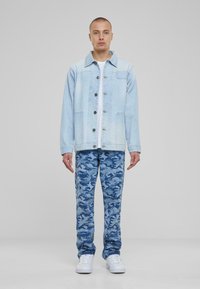 Chaqueta de mezclilla azul claro con cierre de botón y bolsillo en el pecho, combinada con pantalones de camuflaje azul y zapatillas blancas.