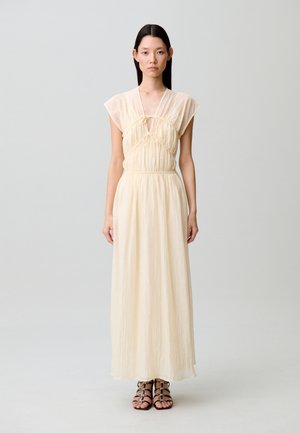 Donna con capelli lunghi e scuri, che indossa un abito senza maniche color crema lungo fino a terra e sandali neri con cinturini, in piedi davanti a uno sfondo chiaro e semplice.