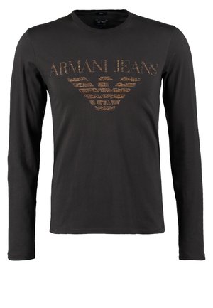 Mężczyzna w ciemnej koszuli z długim rękawem, niebieskich dżinsach, brązowych butach, wielokolorowym szaliku i torbie na ramię idzie do przodu na neutralnym tle.