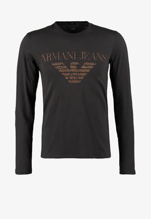 Homme portant une chemise à manches longues foncée, jean bleu, bottes marron, écharpe multicolore et sac bandoulière marchant vers l'avant sur un fond neutre.