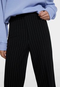 Zwarte broek met streepjes in een gladde stof, hoge taille en wijde pijpen. Gecombineerd met een lichtblauwe trui.