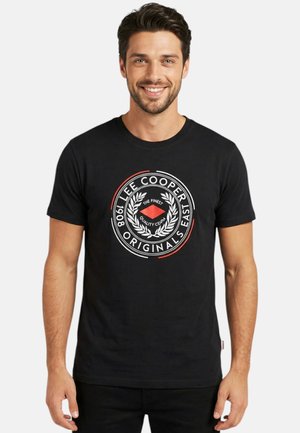 Homme souriant, portant un t-shirt noir "Lee Cooper Originals East 1908" avec un motif circulaire et une couronne de laurier, debout devant un fond uni.