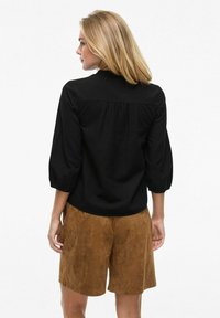 Blusa nera con maniche a tre quarti, tessuto texturizzato e schiena arricciata; abbinata a pantaloni corti in finta pelle scamosciata marrone.