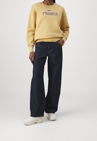 Osoba w musztardowej bluzie z napisem "FRANCJA", w ciemnych szerokich dżinsach, w białych, masywnych trampkach, stojąca z jedną ręką w kieszeni.