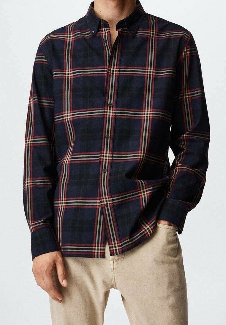 Homme portant une chemise à carreaux bleu marine, rouge et beige à manches longues boutonnée avec un pantalon beige, une main dans la poche, torse visible.