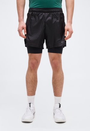 Man wearing black layered running shorts, white athletic socks, and white sneakers, standing against a plain light background.