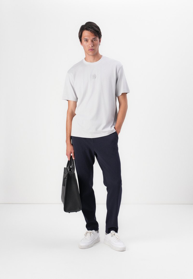 T-shirt grigio chiaro a maniche corte con scollatura rotonda, abbinato a pantaloni blu navy e sneakers bianche. Borsa nera tenuta in una mano.
