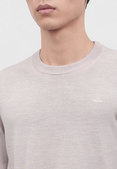 Gros plan d'une personne portant un pull en maille beige clair à col rond avec un logo discret sur la poitrine.