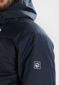 Marinblå vattentät jacka med dragkedja på huvan och en liten vit tassavtryckslogotyp på ärmen. Slät yta och strömlinjeformad design.