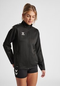 Zwarte zip-up jas met hoge kraag, lange mouwen en een zachte textuur. Bevat een wit logo en een onderscheidend schouderontwerp.