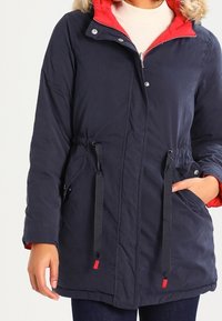 Námořnická modrá parka s červenou podšívkou, umělou kožešinou na kapuci, stahovacími pasem a bočními kapsami. Obsahuje zapínání na patentky.