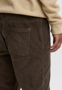 Pantaloni di cotone a coste marroni con finitura testurizzata, dotati di una tasca posteriore e di una tasca laterale, abbinati a una felpa beige.