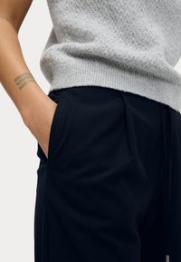Lichtgrijze gebreide trui met een textuurpatroon, en zwarte op maat gemaakte broek met zijzakken en zachte stof, gedragen met de hand in de zak.