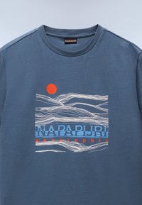 Modrá bavlnená tričko s grafickým dizajnom obsahujúcim vlnité línie v bielej a modrej farbe, oranžové slnko a text "NAPAPIJRI geographic" v tučnom písme.