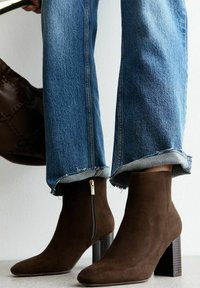 Bruine suède enkelboots met een vierkante neus, zijrits en een gestructureerde blokhak. Gecombineerd met versleten, opgerolde blauwe spijkerbroeken.