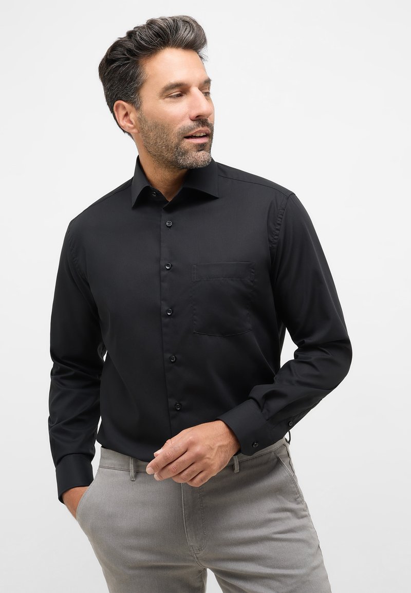 Zwart overhemd met lange mouwen en een spits kraag, met een knoopsluiting aan de voorkant, een enkele borstzak en een gladde textuur, gecombineerd met lichtgrijze broek.