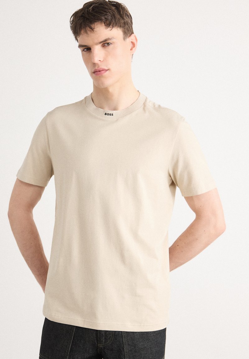 T-shirt beige à manches courtes avec un col rond et un col côtelé. Présente un logo "BOSS" noir au niveau du col. Fabriqué en coton doux.