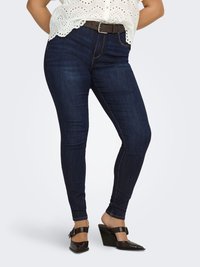 Nainen pukeutuneena tummansinisiin skinny-farkkuihin, ruskeaan vyöhön, valkoiseen silkkipitsipaitaan ja ruskeisiin korkokenkiin, joissa on soljet, seisoo jalat ristissä.