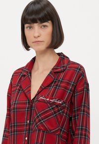 Czerwona koszula piżamowa w kratę z kołnierzykiem, zapinana na guziki i z haftowaną kieszonką z napisem "proszę nie przeszkadzać". Miękki materiał, luźny krój.