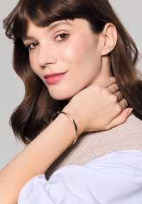 Bransoletka w złotym odcieniu o skręconej fakturze, noszona na nadgarstku. Tło ma jasnoszary odcień, podkreślający blask bransoletki.