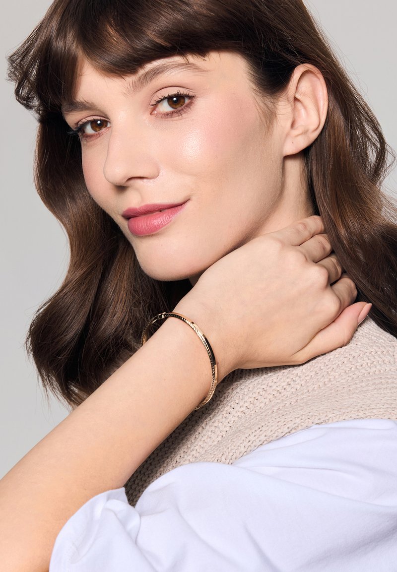 Bransoletka w złotym odcieniu o skręconej fakturze, noszona na nadgarstku. Tło ma jasnoszary odcień, podkreślający blask bransoletki.