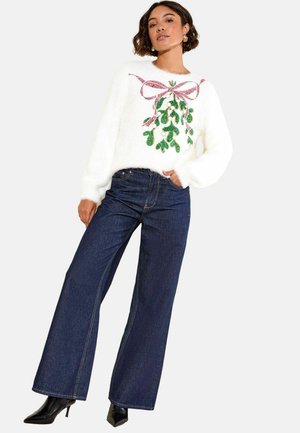 Frau in einem weißen, flauschigen Pullover mit grünem Mistelzweig- und pinkem Schleifenmuster, kombiniert mit dunkelblauen Weitbeine-Jeans und schwarzen Absatzstiefeln.
