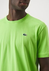 T-shirt in cotone verde con colletto a girocollo, caratterizzato da un piccolo logo di coccodrillo ricamato sul lato sinistro del petto. Texture liscia e maniche corte.