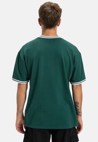 Mężczyzna z krótkimi, brązowymi włosami, odwrócony tyłem, w ciemnozielonej koszulce z krótkim rękawem, z białymi paskami przy kołnierzyku i mankietach.