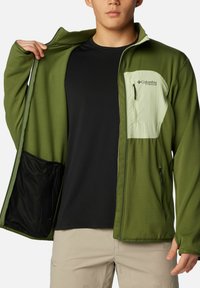Giacca verde con una finitura testurizzata, taschino anteriore verde chiaro e zip frontale. Indossata sopra una maglietta nera e pantaloni beige.