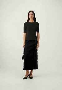 Black, fitted top with short sleeves paired with a long black skirt featuring fringe detailing and a side slit, complemented by black heels.