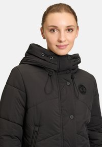 Giacca a vento nera con colletto alto, bottoni frontali e coulisse. Presenta una patch rotonda e tasche laterali con zip.