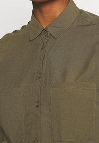 Oliivinvihreä napillinen paita, jossa on kauluksellinen kaulus, kaksi rintataskua ja sileä, kevyesti teksturoitu kangas.