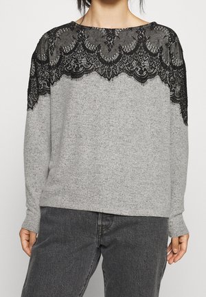 Personne portant un haut gris clair à manches longues avec des détails en dentelle noire sur les épaules et le haut de la poitrine, associé à un pantalon gris foncé.