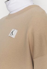 Sweat-shirt beige avec col rond côtelé, texture douce, avec une écusson blanc portant un logo noir sur le côté gauche de la poitrine.