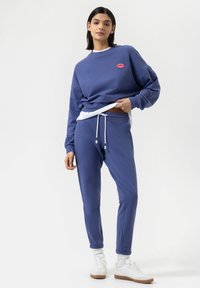 Blauwe katoenen sweatshirt met een rode lip graphic, gecombineerd met bijpassende joggers met een elastische tailleband en witte koordjes, gedragen met witte sneakers.