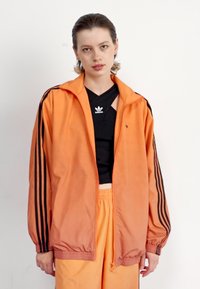 Oransje treningsjakke med svarte striper på ermene, lett materiale, glidelås i front og ribbestrikket kant; brukt over en svart topp og oransje bukser.