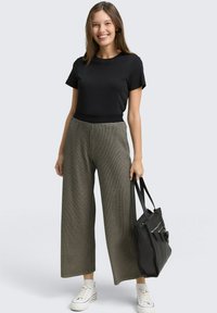 Zwarte geribbelde top met korte mouwen, hoogsluitende grijze broek met wijdnekkige pijpen en een houndstooth patroon, en witte sneakers. Een zwarte schoudertas wordt vastgehouden.