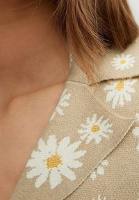 Beżowa tkanina z wzorem białych stokrotek o małych żółtych środkach, z kołnierzykiem i lekką fakturą. Zbliżenie na dekolt.