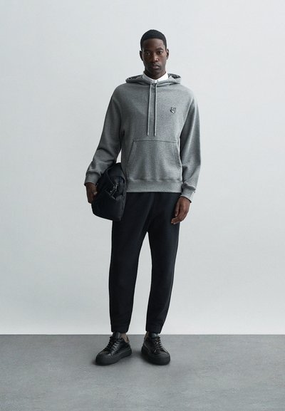Felpa grigia con cappuccio e tasca a marsupio, abbinata a pantaloni neri a gamba stretta e sneakers nere. Il modello tiene in mano una piccola borsa nera.