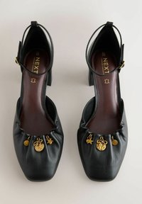 Chaussures à talons hauts en cuir noir avec brides à la cheville et breloques dorées, comprenant une étoile, un cercle, un nœud et un disque martelé sur des bouts froncés.