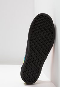 Semelle noire de sneaker avec motif de marche en nid d'abeille, inclinée contre un mur blanc, montrant une partie d'un panneau latéral coloré.