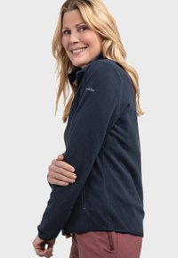 Navy-farbenes Fleecejacket mit hohem Kragen, Seitentaschen und tailliertem Design. Glatte Textur mit einem dezenten Logo-Detail am Ärmels.