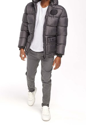 Osoba w szarej kurtce puchowej, białej koszulce, szarych spodniach i białych sneakersach idąca na białym tle.